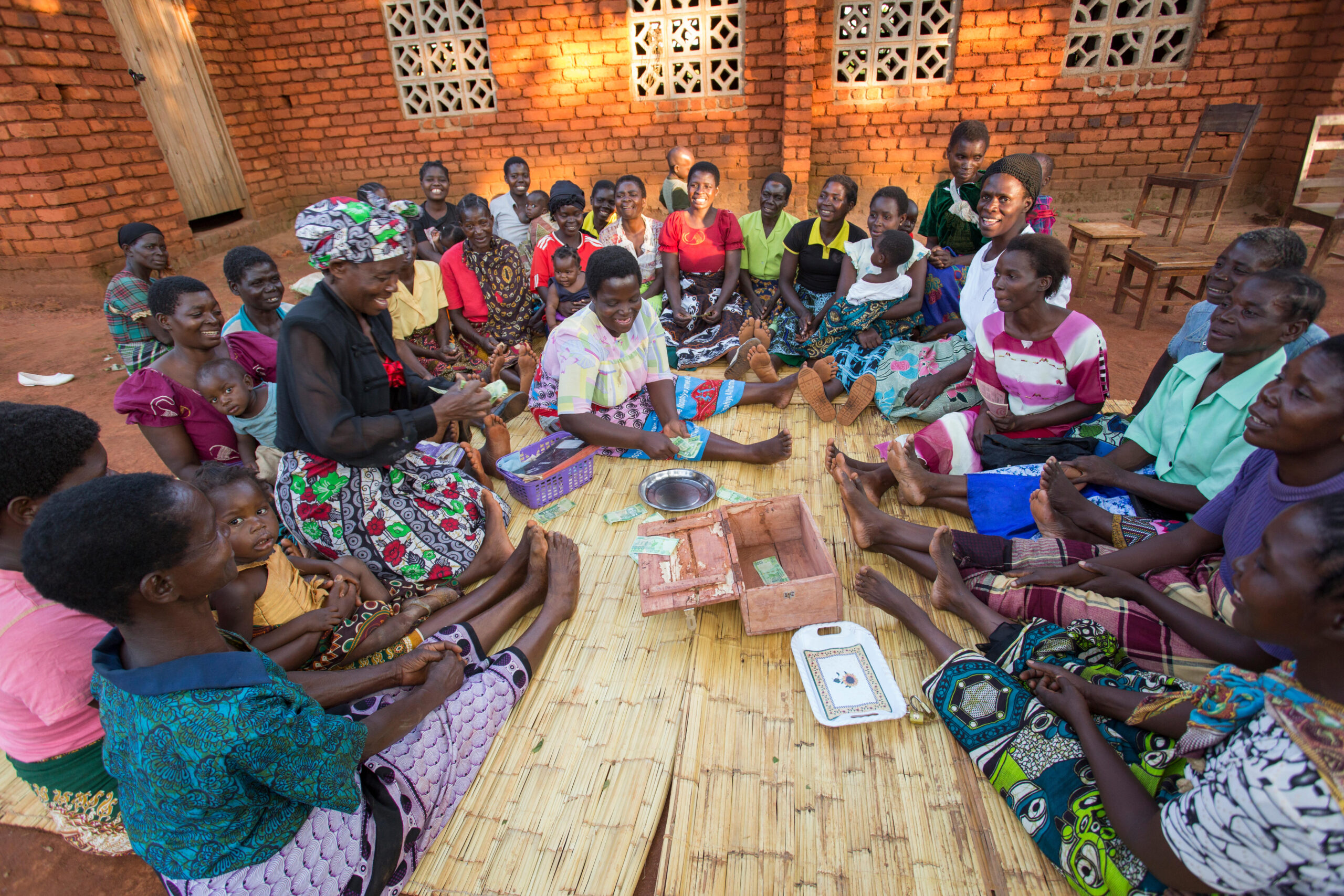 Community giving in southern Africa in action. 20+ women gathered round, sitting on the ground in front of a wooden building that is catching the light of the setting sun.