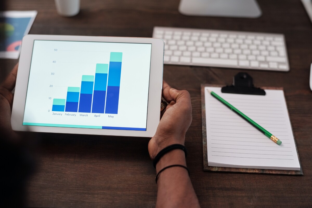 Hands hold a tablet showing a blue shaded barchart, at a desk near a keyboard and a notebook and pen. Photo by Mikael Blomkvist