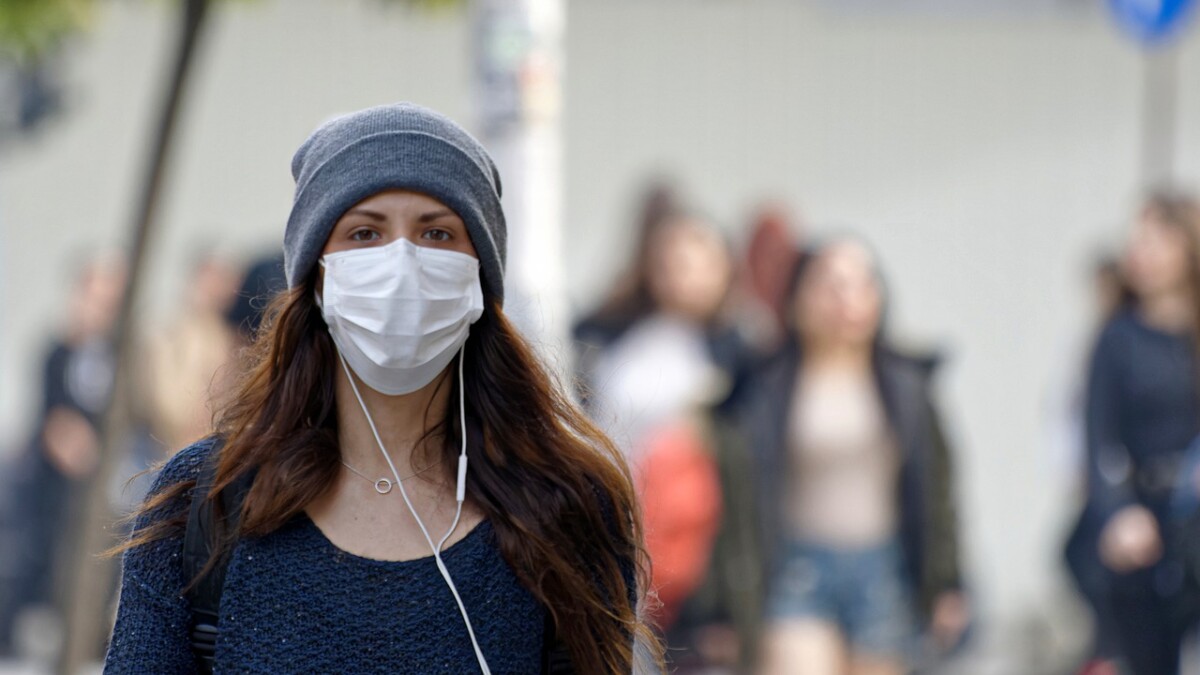 A woman with long brown hair, wearing a beanie and a mask looks at the camera with other people blurred in the background