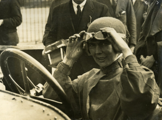 Katherine Martin, putting no goggles, seated at the wheel of a racing car. A black and white image.